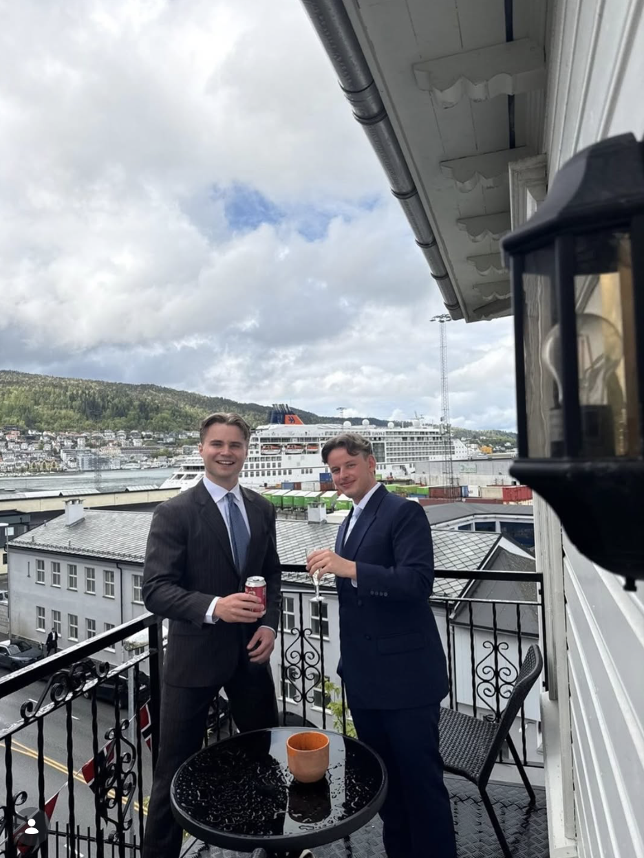 To menn i dresser på balkong med utsikt over havn og by